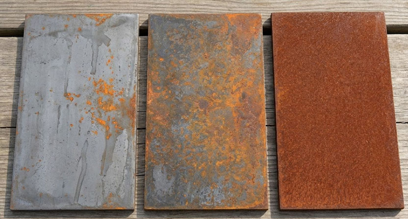 Drei nebeneinander gestellte Cortenstahl-Platten in unterschiedlichen Rostierungsstadien, von blank grau über fleckigen Flugrost bis zu geschlossener rotbrauner Patina, im weichen Tageslicht fotografiert