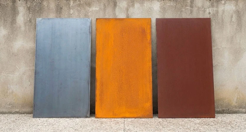 Drei flache Cortenstahlplatten lehnen nebeneinander an einer Wand: links noch blaugrau roh, mittig hell orange nach wenigen Monaten, rechts tief rotbraun nach Jahren
