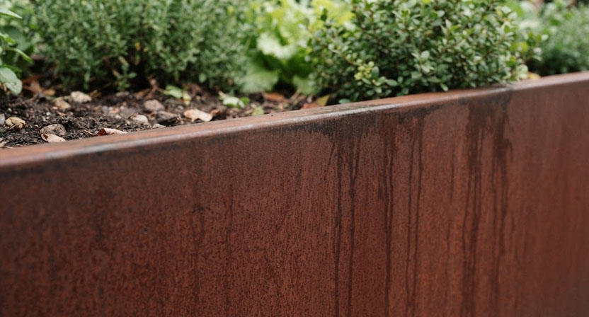 Nahaufnahme einer Cortenstahl-Hochbeetwand von schräg oben: tiefe rotbraune Patina mit feiner, samtig matter Textur und dunkleren Regenlaufspuren