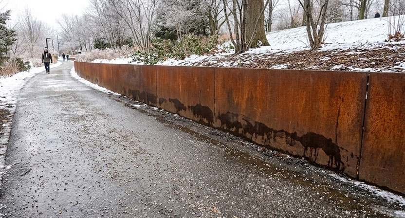 Ein gestreuter Gehweg direkt neben einer niedrigen Cortenstahl-Stützmauer, an deren unterem Rand sich dunklere, unregelmäßige Verfärbungen durch Spritzwasser zeigen.